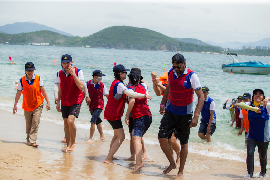 Nhóm nhân viên mặc áo team màu vàng và cam cùng tham gia hoạt động team building trên bãi biển, minh họa cho hiệu quả khi chọn đơn vị tổ chức chuyên nghiệp và sáng tạo.