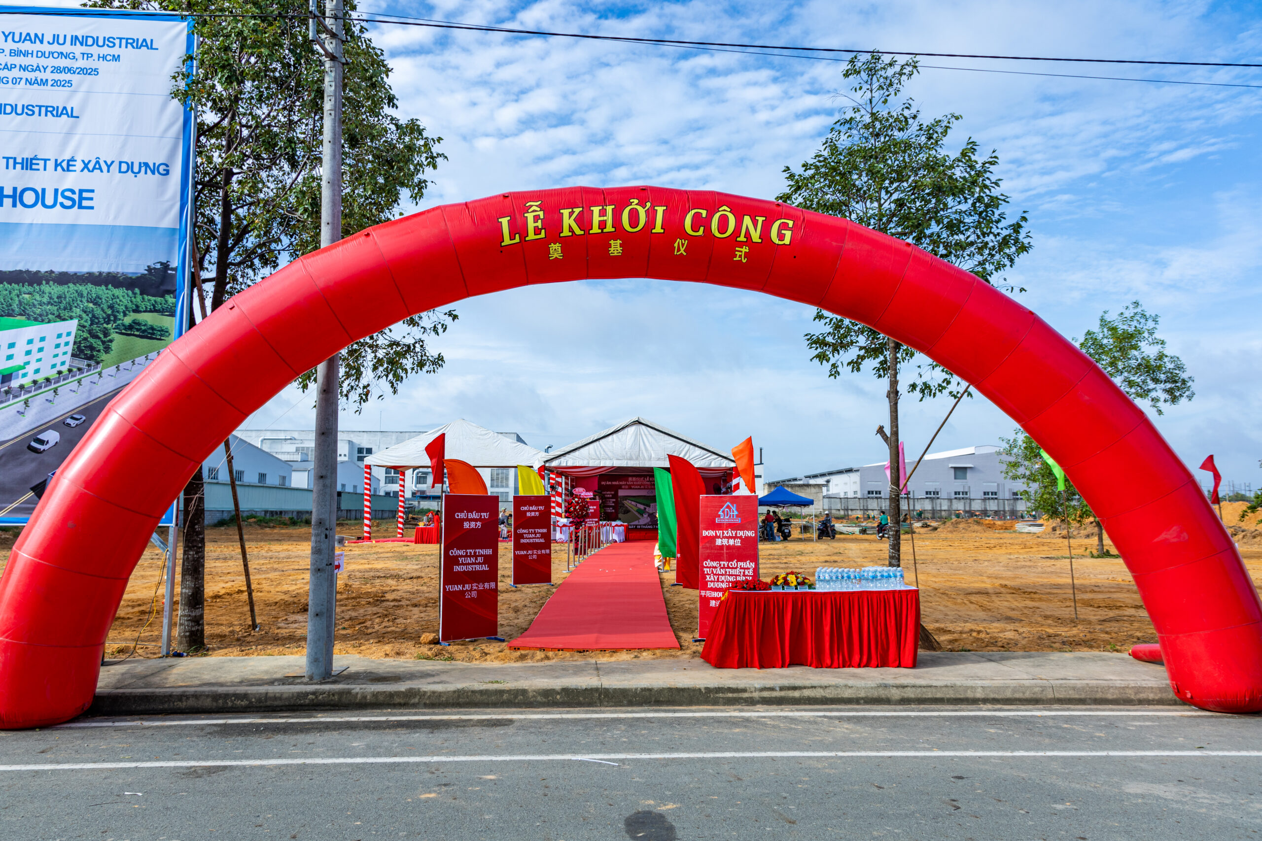 alt= "Không gian lễ khởi công dự án xây dựng với cổng chào đỏ, thảm đỏ và khu vực đón tiếp khách mời trang trọng."